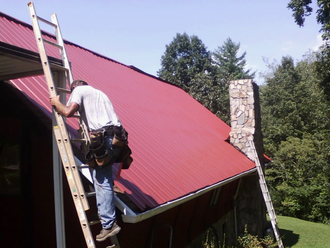 Roofer on a ladder beside a red metal roof and stone chimney — Soffit & Fascia Repair by Roofing Brothers in Seguin